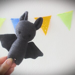 Puede incluir: Un juguete de peluche de murci&eacute;lago de fieltro gris con orejas grandes y redondeadas y una cara sonriente simple. El murci&eacute;lago est&aacute; colgado de una cuerda de banderas de papel de colores.