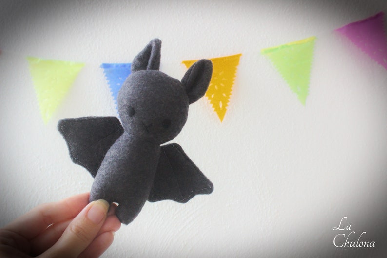 Puede incluir: Un juguete de peluche de murci&eacute;lago de fieltro gris con orejas grandes y redondeadas y una cara sonriente simple. El murci&eacute;lago est&aacute; colgado de una cuerda de banderas de papel de colores.