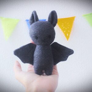 Puede incluir: Un juguete de peluche de murci&eacute;lago de fieltro gris con ojos grandes y negros y una peque&ntilde;a sonrisa cosida. El murci&eacute;lago est&aacute; sentado en una mano sobre un fondo blanco.