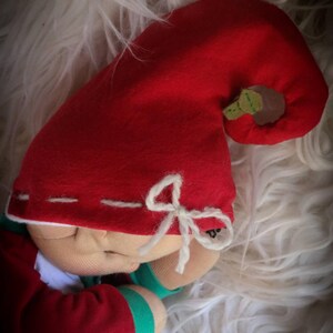 May include: A handmade doll with a red felt hat and matching red and green outfit. The hat features a small green leaf and a white bow. The doll is resting on a fluffy white surface.