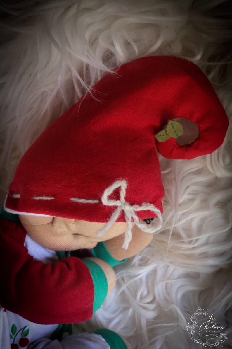 May include: A handmade doll with a red felt hat and matching red and green outfit. The hat features a small green leaf and a white bow. The doll is resting on a fluffy white surface.