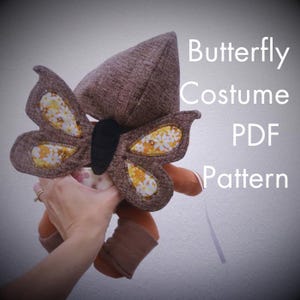 May include: A brown fabric butterfly costume with a pointed hood and wings featuring yellow and white floral patterned inserts. The costume has a black central body section. The text Butterfly Costume PDF Pattern is visible on the right side.