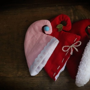 May include: Three elf hats in pink and red with white trim and accents. One hat is pink with a blue pom-pom and white lace trim. The other two are red, one with a white bow and the other with a white fluffy trim.