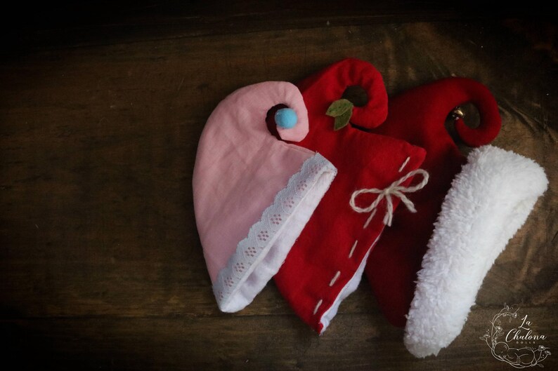May include: Three elf hats in pink and red with white trim and accents. One hat is pink with a blue pom-pom and white lace trim. The other two are red, one with a white bow and the other with a white fluffy trim.