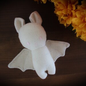 Puede incluir: Un murci&eacute;lago de peluche blanco con ojos rosas y una sonrisa cosida. El murci&eacute;lago tiene alas grandes y est&aacute; acostado sobre una superficie de madera marr&oacute;n.