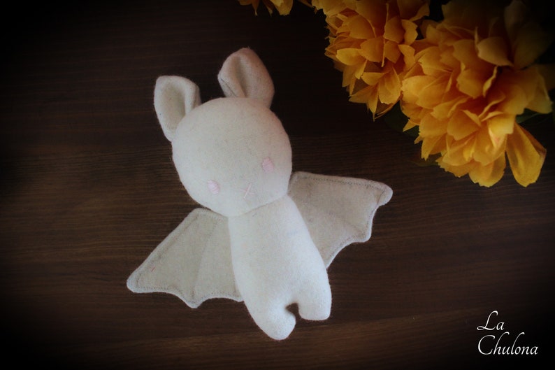 Puede incluir: Un murci&eacute;lago de peluche blanco con ojos rosas y una sonrisa cosida. El murci&eacute;lago tiene alas grandes y est&aacute; acostado sobre una superficie de madera marr&oacute;n.