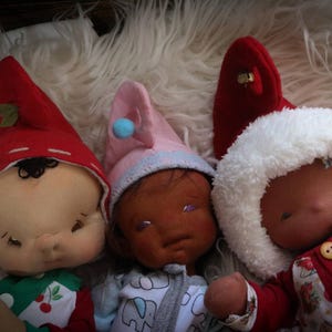 May include: Three handmade cloth dolls with closed eyes, each wearing a different hat. One doll wears a red hat with a green leaf, another a pink hat with a blue pom-pom, and the third a white fluffy hat. The dolls are lying on a white, fluffy surface.