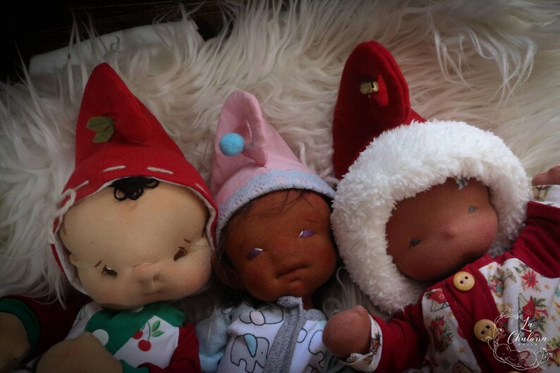 May include: Three handmade cloth dolls with closed eyes, each wearing a different hat. One doll wears a red hat with a green leaf, another a pink hat with a blue pom-pom, and the third a white fluffy hat. The dolls are lying on a white, fluffy surface.
