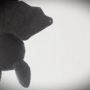 Puede incluir: Un juguete de peluche de murci&eacute;lago negro con una textura suave y peluda. El murci&eacute;lago est&aacute; colgado boca abajo con las alas extendidas.