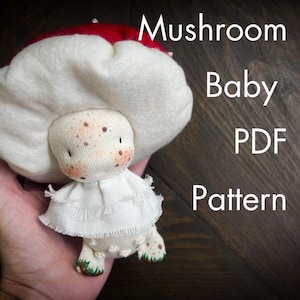 May include: A handmade mushroom baby doll with a white cap and a red top. The doll has a white dress and brown spots on its face. The text "Mushroom Baby PDF Pattern" is visible.