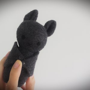 Puede incluir: Un juguete de murci&eacute;lago de peluche gris con una cara linda. El murci&eacute;lago est&aacute; doblado por la mitad y sostenido por una mano.