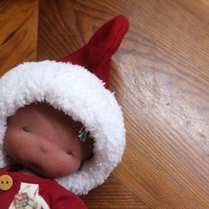 May include: A handmade doll wearing a red outfit and a white fur-trimmed hat. The doll's face is a soft pink colour, and it has small, embroidered eyes. The doll is lying on a wooden surface.