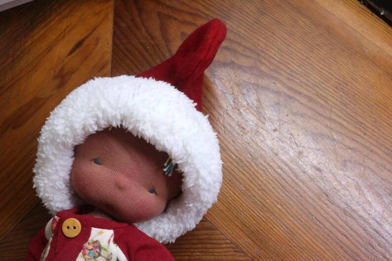 May include: A handmade doll wearing a red outfit and a white fur-trimmed hat. The doll's face is a soft pink colour, and it has small, embroidered eyes. The doll is lying on a wooden surface.