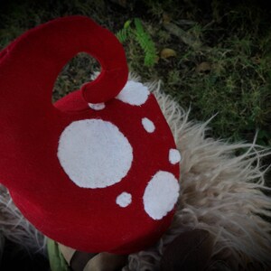 May include: A red mushroom hat with white spots. The hat has a curved stem and is made of a soft material. It is likely a costume piece or a whimsical accessory.