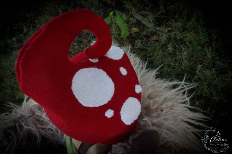 May include: A red mushroom hat with white spots. The hat has a curved stem and is made of a soft material. It is likely a costume piece or a whimsical accessory.