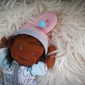 May include: A handmade doll with brown skin, wearing a pink nightcap with a blue pom-pom and a light blue and white onesie with elephant print. The doll is lying on a fluffy white surface.