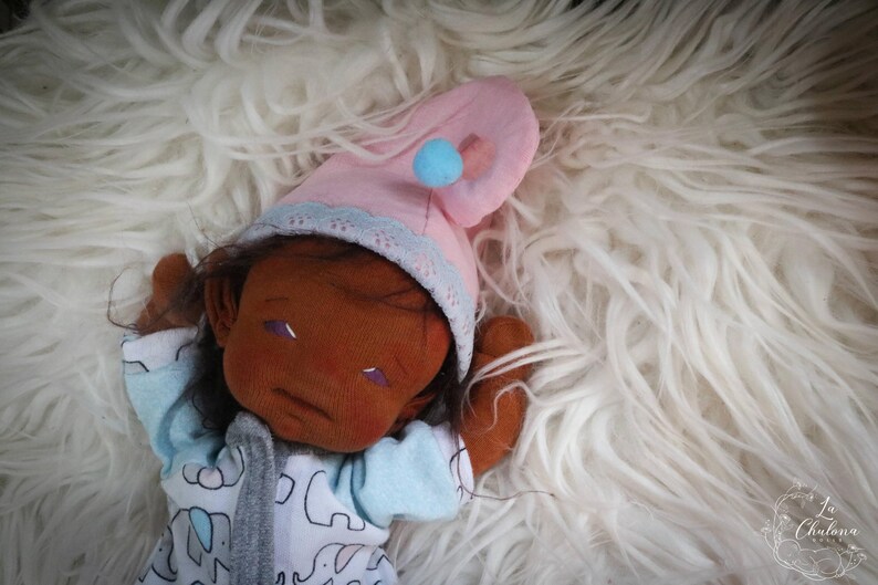 May include: A handmade doll with brown skin, wearing a pink nightcap with a blue pom-pom and a light blue and white onesie with elephant print. The doll is lying on a fluffy white surface.