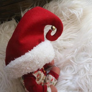 May include: A red felt elf hat with a white fluffy trim and a small gold bell. The hat is on a white fluffy surface. The baby is wearing a red outfit with a floral pattern.