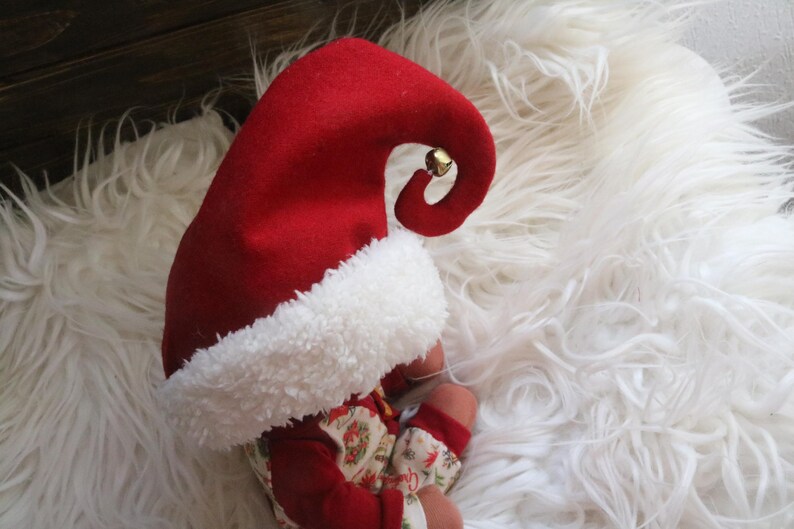 May include: A red felt elf hat with a white fluffy trim and a small gold bell. The hat is on a white fluffy surface. The baby is wearing a red outfit with a floral pattern.
