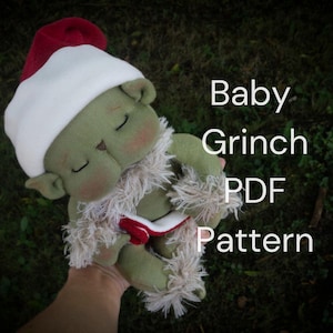 Könnte beinhalten: Eine handgefertigte grüne Plüschpuppe mit roter und weißer Weihnachtsmütze und einem flauschigen weißen Kragen. Die Puppe liegt mit geschlossenen Augen. Der Text auf dem Bild lautet "Baby Grinch PDF Pattern."