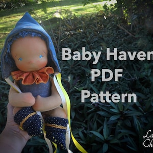 May include: A handmade cloth doll wearing a blue hooded outfit with a yellow ribbon. The doll is holding its hands together and has blue eyes. The background is a green leafy bush. The text "Baby Haven PDF Pattern" is written in white on a black background.