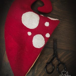 May include: A red felt mushroom hat with white spots and a pair of black scissors. The hat has a curved top and a triangular base. The scissors have ornate handles. The background is a dark wood surface.