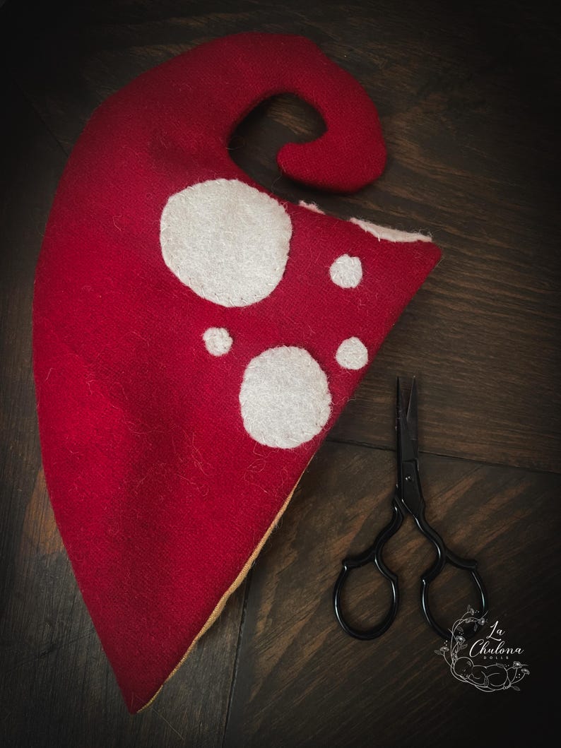 May include: A red felt mushroom hat with white spots and a pair of black scissors. The hat has a curved top and a triangular base. The scissors have ornate handles. The background is a dark wood surface.