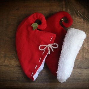 May include: Two red elf hats with white trim and a white fluffy stocking. One hat has a small green leaf and the other has a small gold bell. The hats are tied with a white string and sit on a wooden surface.