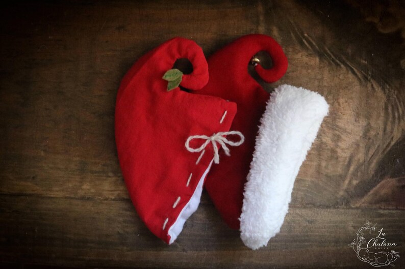 May include: Two red elf hats with white trim and a white fluffy stocking. One hat has a small green leaf and the other has a small gold bell. The hats are tied with a white string and sit on a wooden surface.