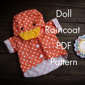 May include: An orange doll raincoat with white polka dots and a duck-shaped hood. The raincoat has a yellow beak and black eyes. The text "Doll Raincoat PDF Pattern" is visible on the image. The raincoat has white buttons and a plaid lining.