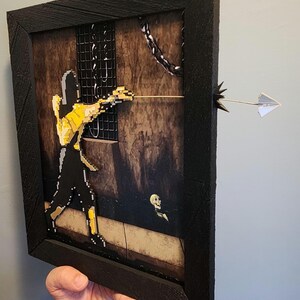Può includere: Opera d'arte incorniciata con una figura pixelata in nero e giallo che mira una freccia. La freccia si estende dalla cornice, con un aeroplanino di carta sulla punta. Lo sfondo raffigura una scena di dungeon con catene e un teschio.