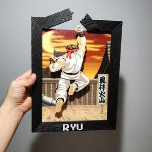 Könnte beinhalten: Ein geschichtetes 3D-Wandkunstwerk, das den Charakter Ryu aus Street Fighter in einer dynamischen Pose zeigt. Der schwarze Rahmen hat eine Holzmaserung und eine abgebrochene Oberkante. Die Szene umfasst einen Sonnenuntergangshintergrund, eine Holzplattform und ein Schild mit japanischen Schriftzeichen. Der Name RYU ist unten in weißen, verpixelten Buchstaben aufgedruckt.