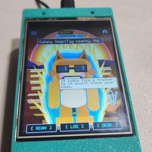 May include: A teal handheld electronic device with a screen displaying a pixelated character wearing sunglasses, surrounded by a blue aura. The screen text reads: Galaxy SmartTag nearby. Hm. It looks like a tracker. Is it yours? Check your keys.