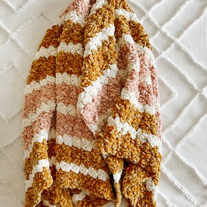 May include: A soft, striped throw blanket in shades of mustard yellow, pink, and white. The blanket is made of a plush, velvety material and is folded in a triangular shape.