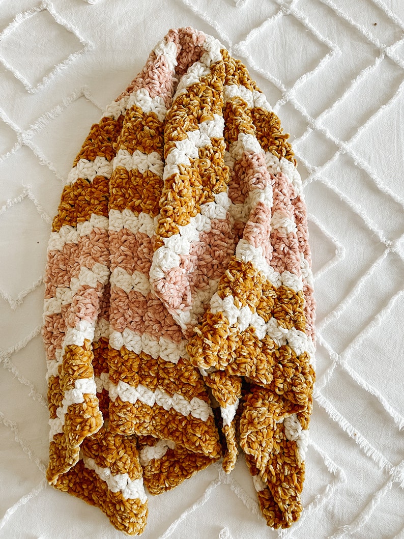 May include: A soft, striped throw blanket in shades of mustard yellow, pink, and white. The blanket is made of a plush, velvety material and is folded in a triangular shape.