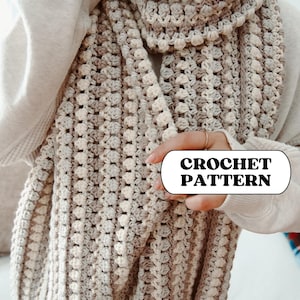 May include: A chunky, textured crochet scarf in neutral tones of cream, beige, and gray. The scarf features a bobble stitch pattern and is wrapped around the neck. The text "CROCHET PATTERN" is visible on the image.