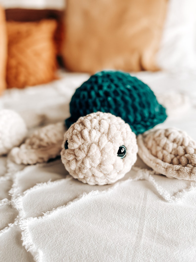 Puede incluir: Una tortuga de peluche tejida a crochet con una concha verde turquesa y un cuerpo de color crema. La tortuga tiene ojos negros y est&aacute; tumbada sobre una colcha blanca.