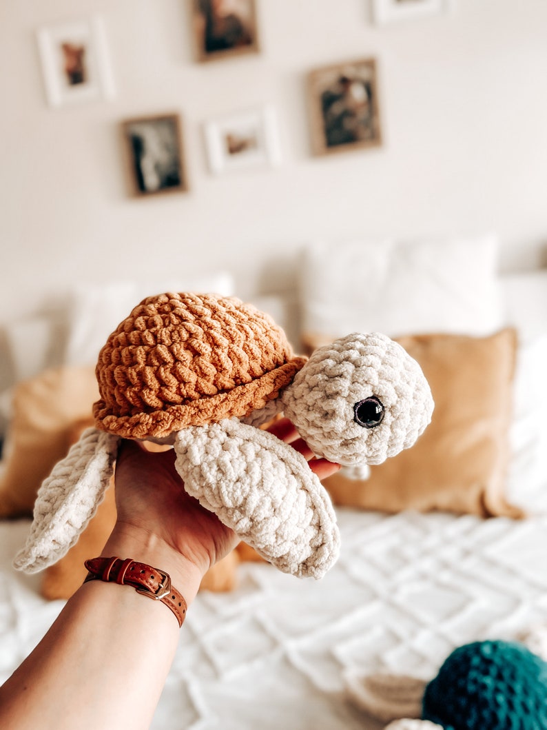 Puede incluir: Un juguete de tortuga de crochet con una concha marr&oacute;n y un cuerpo blanco. El juguete tiene ojos negros y se sostiene en una mano.