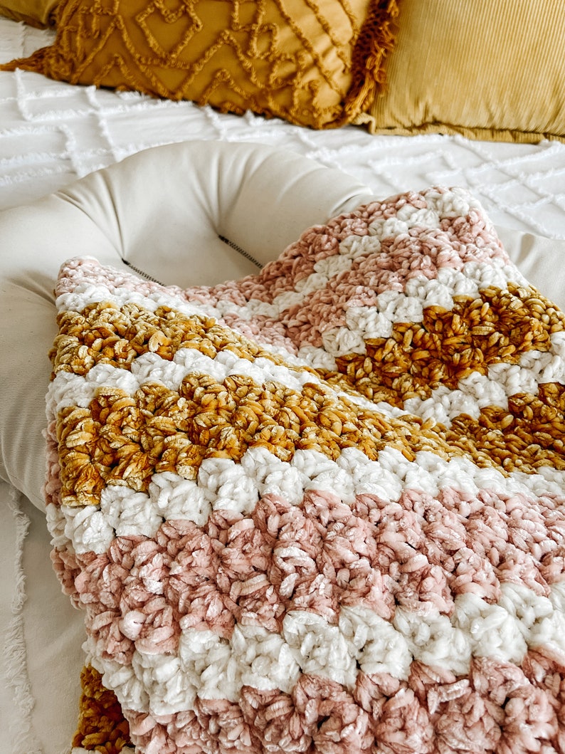 May include: A soft, plush blanket with stripes of pink, white, and mustard yellow. The blanket is made of a chenille yarn and has a fuzzy texture.