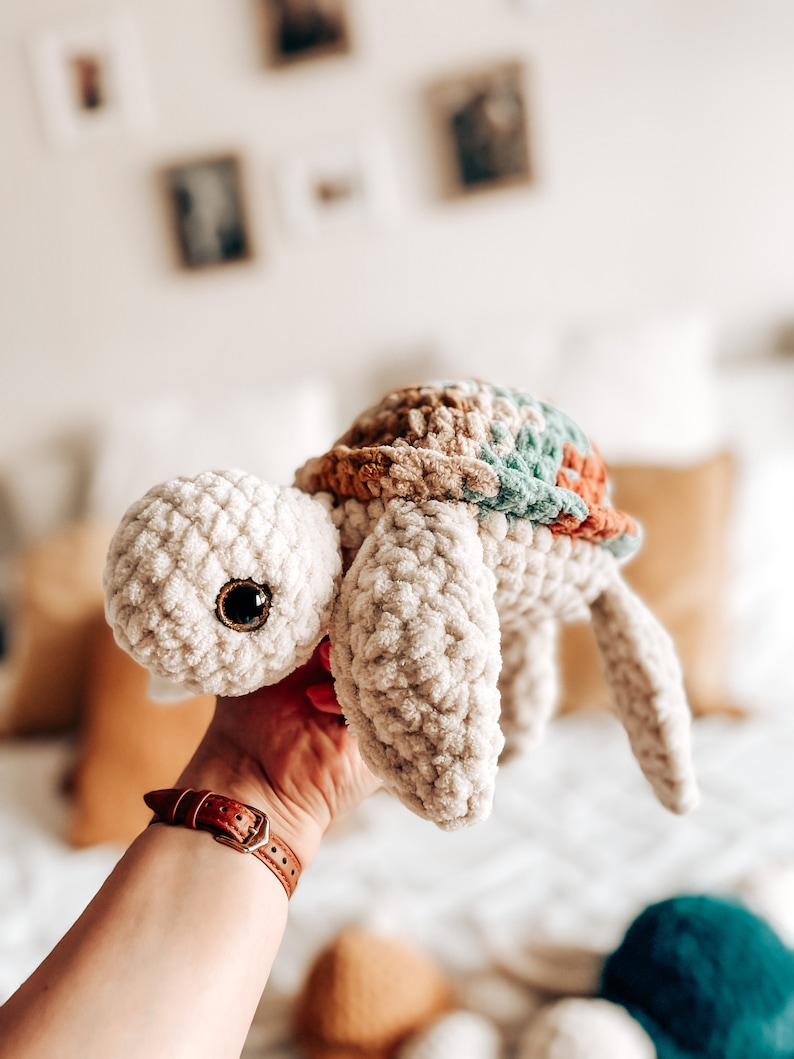 Puede incluir: Un juguete de peluche de tortuga de crochet con un cuerpo beige, un caparaz&oacute;n marr&oacute;n y verde y grandes ojos marrones.