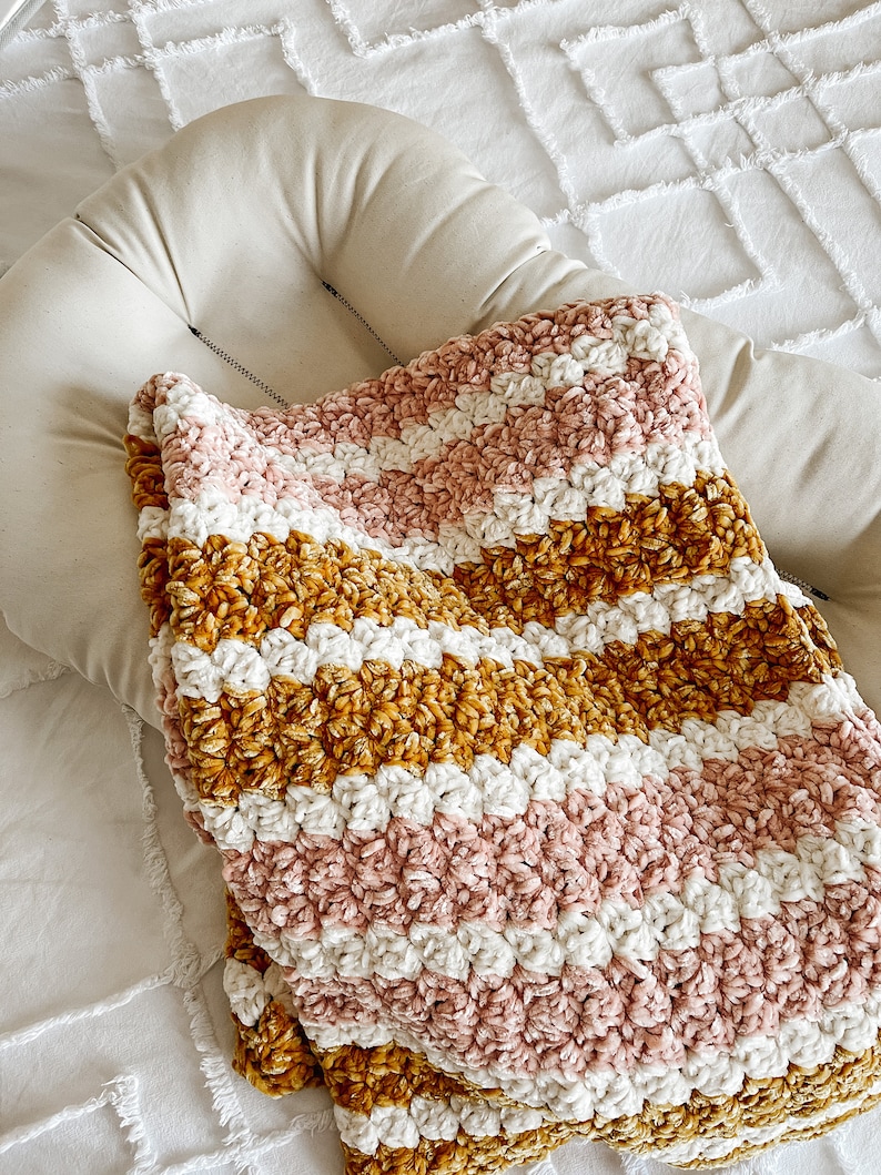 May include: A soft, striped crocheted blanket in shades of pink, white, and mustard yellow. The blanket is made with a fluffy yarn and has a textured, chunky look.