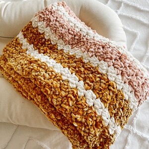 May include: A soft, striped throw blanket in shades of mustard yellow, white, and blush pink. The blanket is made of a fuzzy, textured yarn and is folded on a white surface.