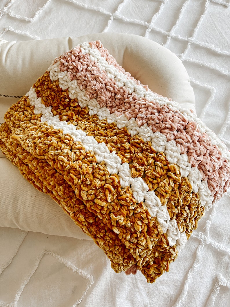 May include: A soft, striped throw blanket in shades of mustard yellow, white, and blush pink. The blanket is made of a fuzzy, textured yarn and is folded on a white surface.