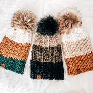 May include: Three knitted hats with different colour combinations. The hats are made with chunky yarn and have a fluffy pompom on top. The colours include beige, brown, green, and black.