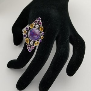 May include: A silver ring with a large, round, purple gemstone at the center, surrounded by smaller yellow, purple, and clear gemstones. The ring is displayed on a black hand-shaped jewelry display.
