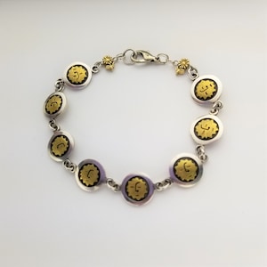 May include: A silver bracelet with gold accents features a repeating pattern of circular charms with a sun design. Each charm has a raised, stylized sun with a face inside.