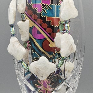 May include: A white gemstone necklace with iridescent green and blue beads. The necklace is displayed in a crystal glass. The background is a colorful patterned fabric.