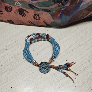 May include: A multi-strand bracelet with blue, brown, and silver beads. The bracelet has a brown leather tassel and a metal charm with a tree design.