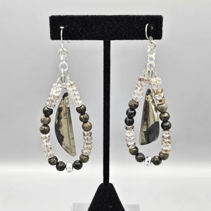 May include: A pair of dangle earrings with a silver chain and a teardrop-shaped design. The earrings feature a large, flat, dark gray stone with gold flecks, surrounded by black, gold, and clear beads.