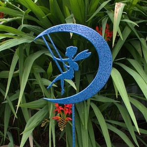 May include: A blue metal garden stake with a crescent moon design. A fairy silhouette is on the moon, swinging on a rope. The stake is in a garden setting with green grass and red flowers.